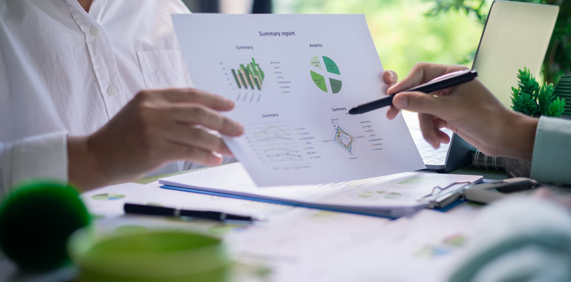 Two people at a desk reviewing a printed summary report of green charts and graphs, one pointing with a pen.